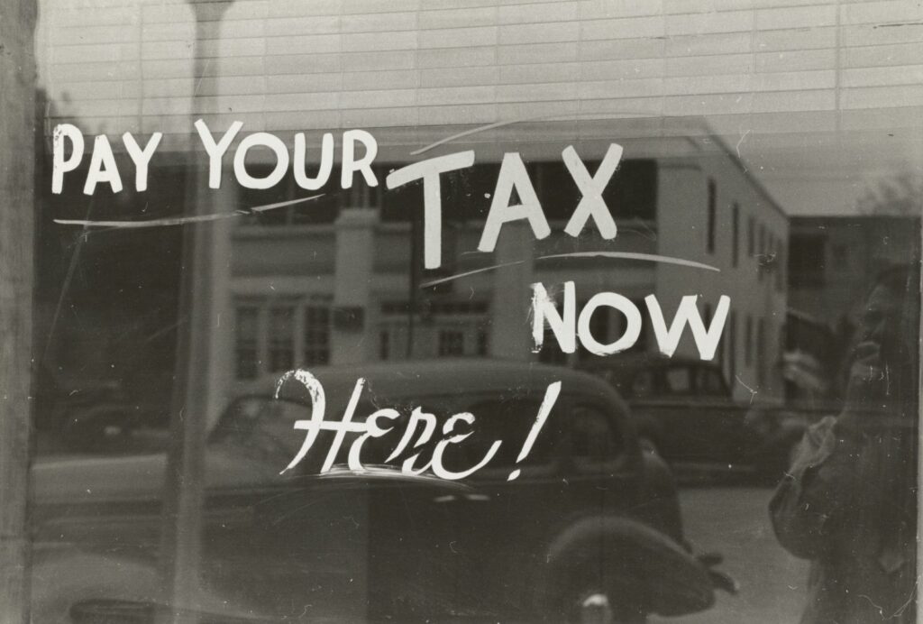 Vintage black-and-white photo of a shop window sign reading Pay Your Tax Now Here